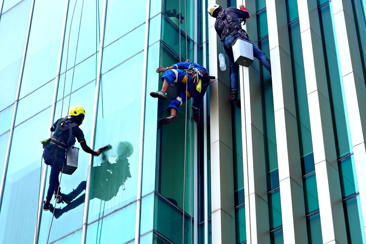 Trois laveurs de vitres en suspension sur des cordes nettoient, de jour, la façade vitrée d’un immeuble moderne.